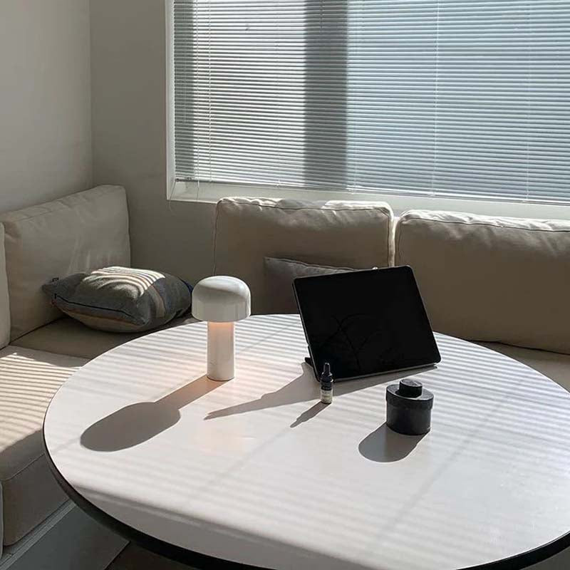A rechargeable Mushroom LED Table Lamp sits on a modern round white coffee table in a minimalist living room, complemented by a black tablet. A cream-colored corner sofa and a window with horizontal blinds are in the background.