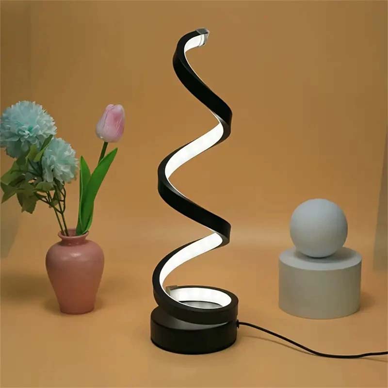 A modern spiral-shaped LED table lamp with a glossy black frame and white illuminated light band, standing on a round black base. The lamp is styled on a warm beige surface next to a pink vase with flowers and a small gray sphere lamp on a cylinder pedestal.