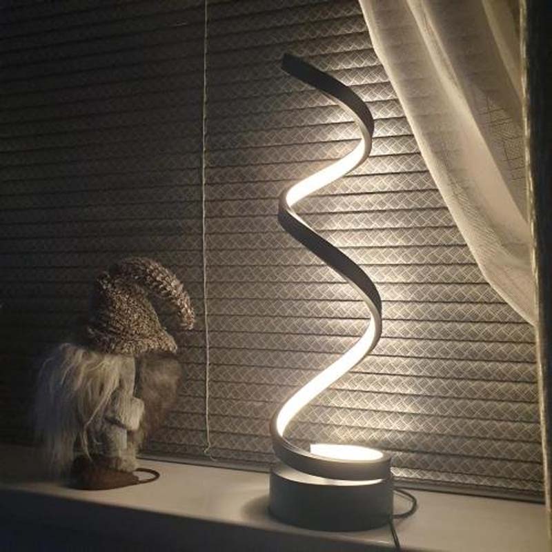 A modern spiral LED table lamp with a black base and a glowing white curved light bar, shown on a windowsill next to a decorative gnome figurine in front of window blinds.