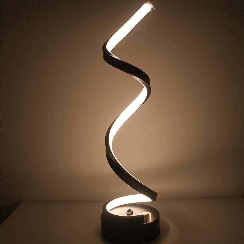 A modern spiral LED table lamp with a black frame and a circular base, shown illuminated against a warm, dimly lit background.