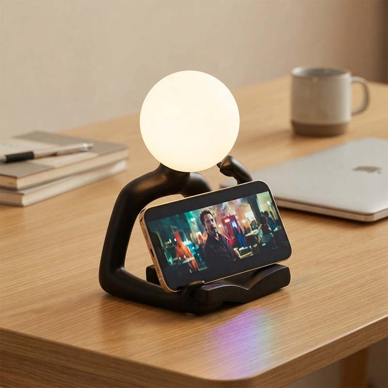 Thinker Statue LED Table Lamp sits on a wooden table, used as a smartphone stand, with a mug and laptop in the background.