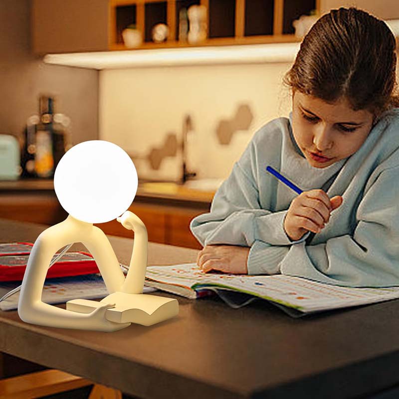 The photo shows a child in a light blue sweatshirt writing at a table, next to a Thinker Statue LED Table Lamp.