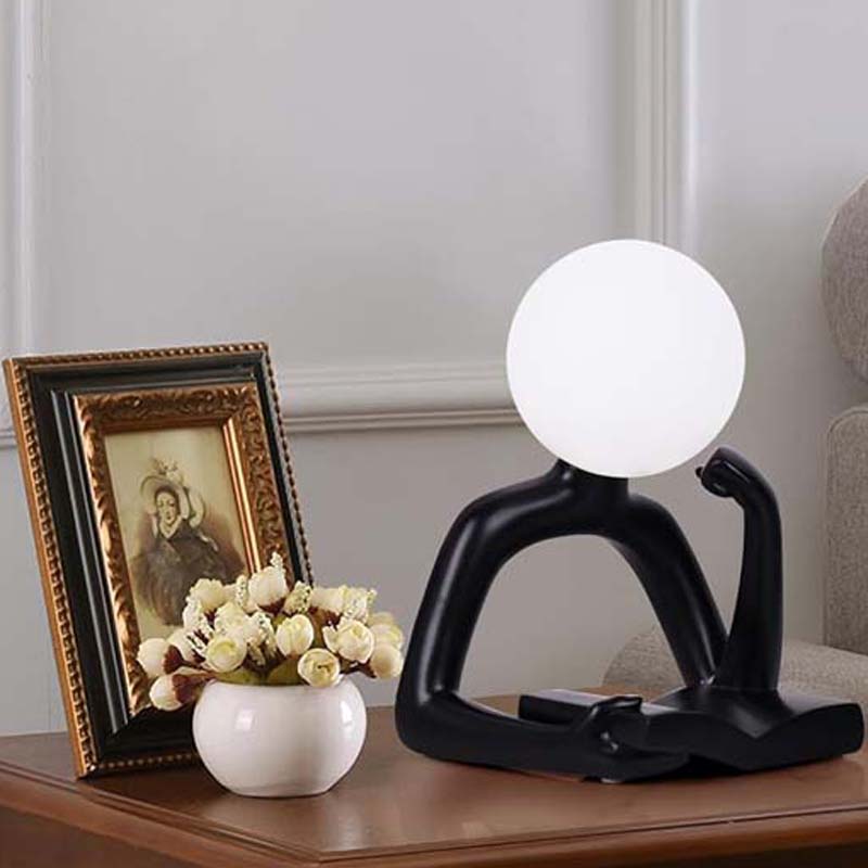 Thinker Statue LED Table Lamp sits on a wooden table next to a framed painting and a vase of white roses.