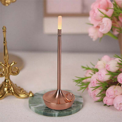 Decorative candle holder with marble base and gold accents on a surface with flowers and a lamp in the background.