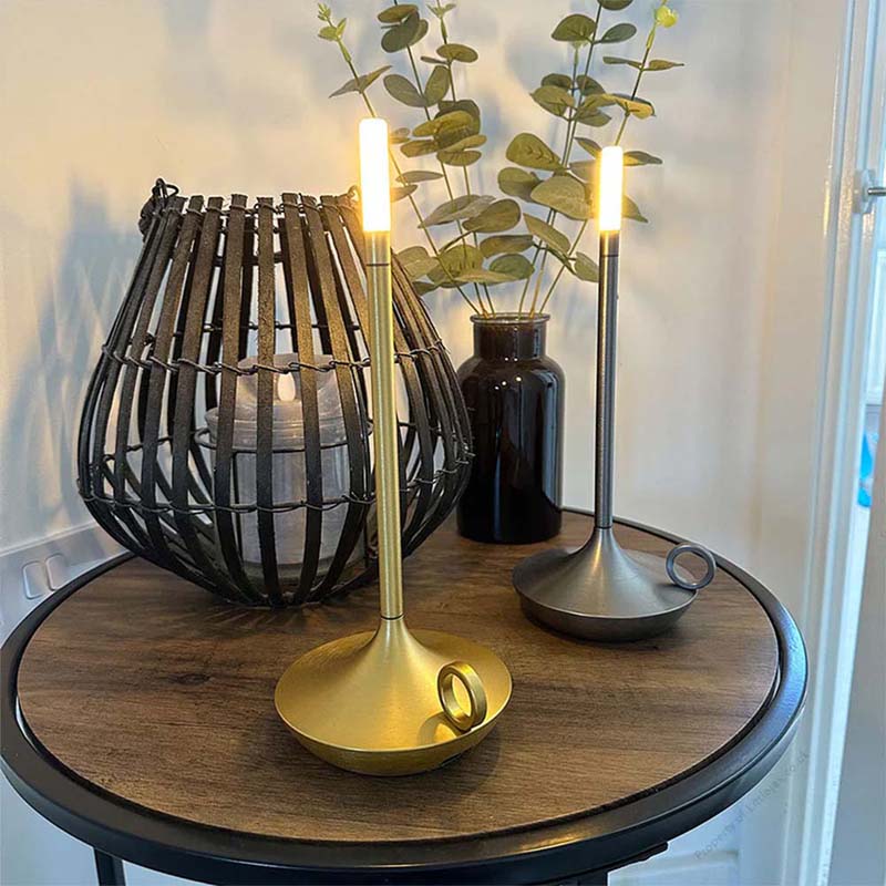Decorative table setting with a gold candle holder, black woven lantern, and vase on a wooden table.