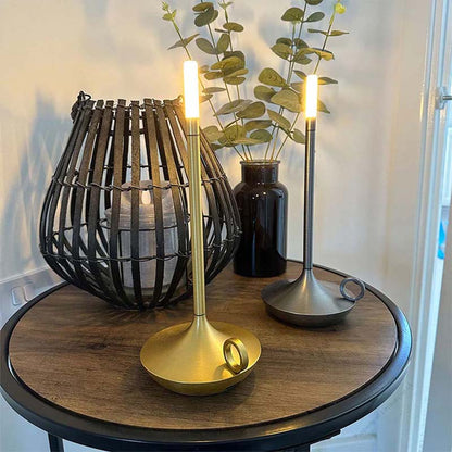 Decorative table setting with a gold candle holder, black woven lantern, and vase on a wooden table.