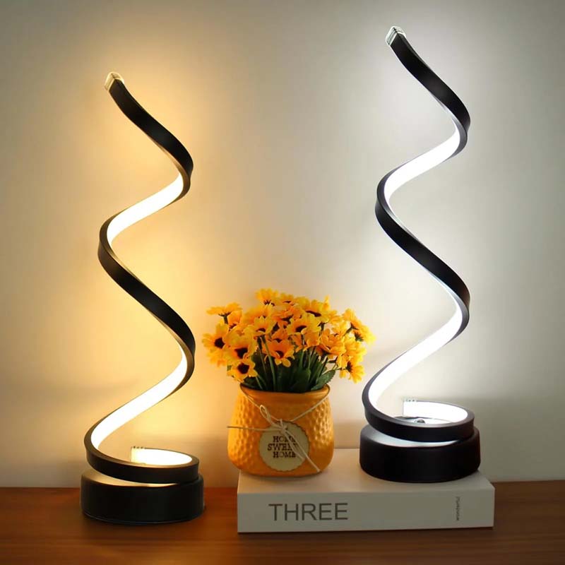 Two modern spiral LED table lamps with black bases and glowing white light, displayed on a wooden surface with a yellow flower pot and a white book in the background.