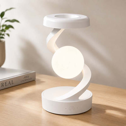 The Saturn Helix – LED Desk Lamp with Wireless Charging is shown glowing on a wooden tabletop with a blurred image of a plant and book in the background.
