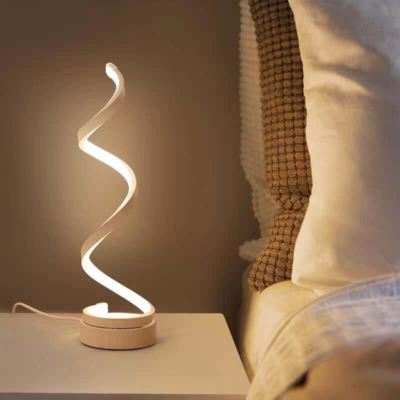 A modern spiral-shaped LED night light with a warm white glow, sitting on a bedside table next to a bed with pillows.