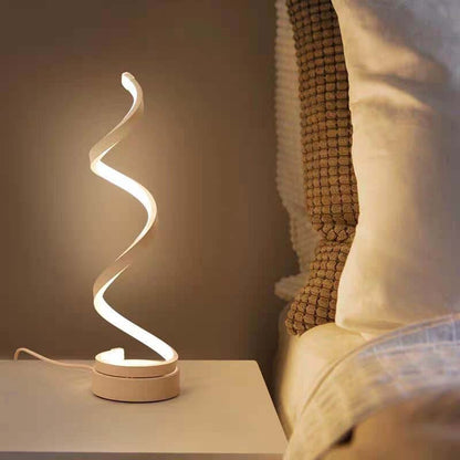 A modern spiral-shaped LED night light with a warm white glow, sitting on a bedside table next to a bed with pillows.