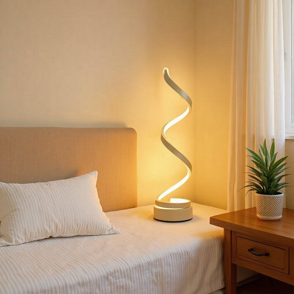 A modern spiral LED table lamp with a white base and curved arm, glowing with a warm yellow light, placed on a bed next to a wooden nightstand with a potted plant.