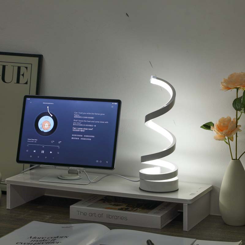 A modern spiral-shaped LED desk lamp glowing on a white monitor riser in a home office setting, with a computer monitor, books, and a vase of flowers nearby.