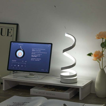 A modern spiral-shaped LED desk lamp glowing on a white monitor riser in a home office setting, with a computer monitor, books, and a vase of flowers nearby.