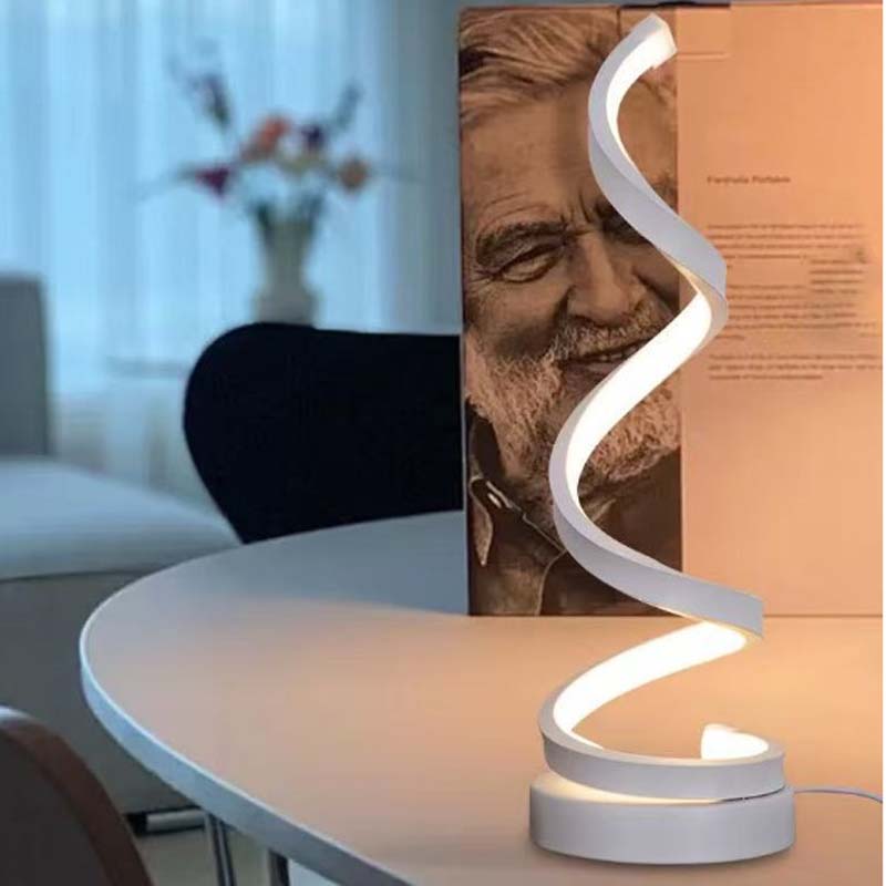 A modern spiral-shaped LED desk lamp with a white curved body, illuminated and placed on a light-colored table in a home office setting next to a book.