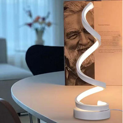 A modern spiral-shaped LED desk lamp with a white curved body, illuminated and placed on a light-colored table in a home office setting next to a book.