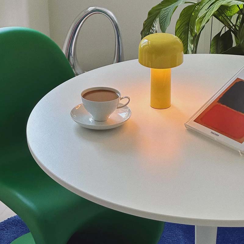 A mushroom-shaped yellow table lamp glowing on a white round table, next to a cup of coffee, a green chair, and a book.